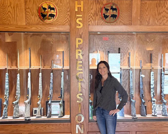 The image shows a person standing in front of a display of rifles at H-S Precision. The display is housed in a wooden cabinet with various rifles lined up. There are decorative signs featuring animal silhouettes above the cabinet.