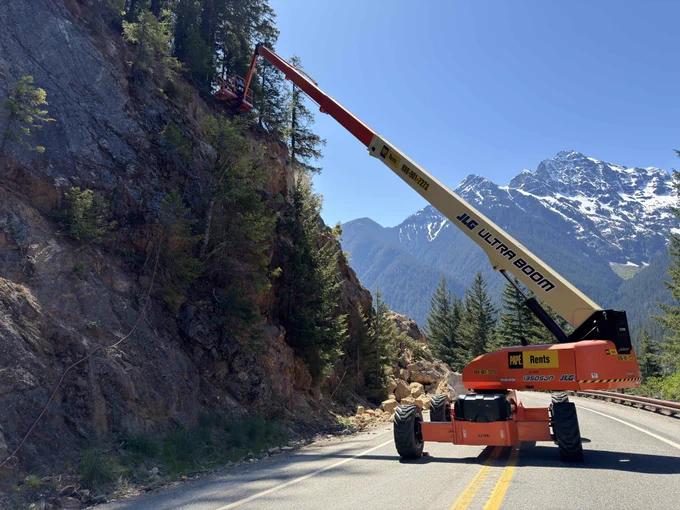 An orange and white construction left sits at the base of a slope with a pile of rocks behind it. Crew members inspect the top of the slope. A mountain vista is in the background on a sunny day.