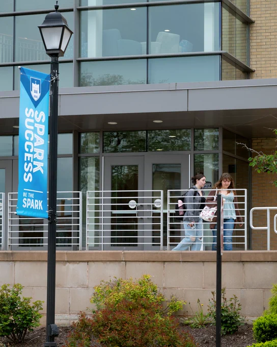 Two students walk outside of Aquinas Hall. A flag pole is seen to the left of the image with a banner that reads: "Spark Success" with "Be the Spark" underneath it.