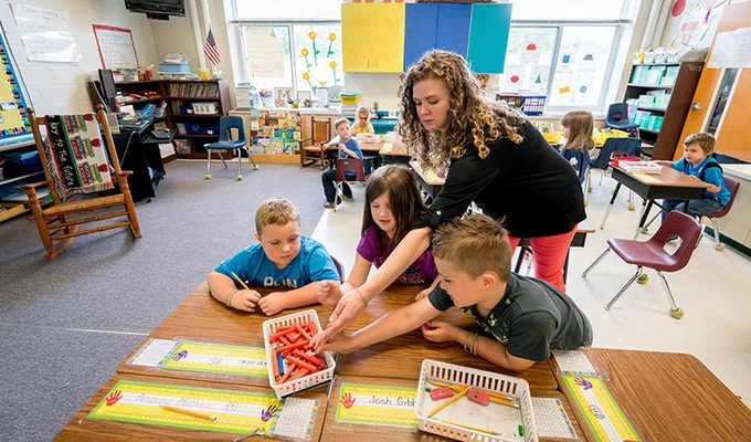 Teacher helping young students select crayons at a classroom table during an art activity.