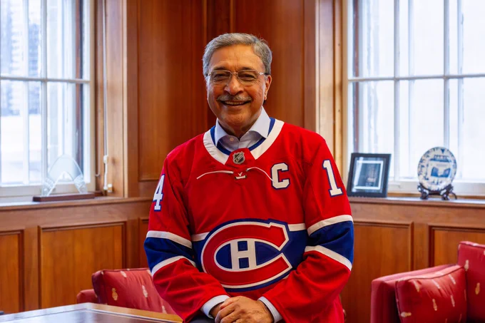 President Saini stands in a wood-paneled office wearing a red Montreal Canadiens hockey jersey, with tall windows and red chairs in the background.