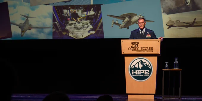 A military officer in uniform speaks at a podium with the Ogden Eccles Conference Center and HIPE logos, with aviation images projected behind him.