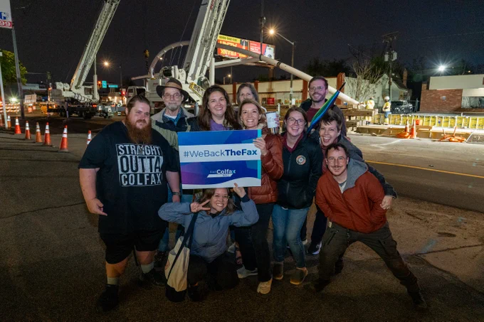 Smiling group at night holding "#WeBackTheFax" sign in front of Colfax street construction and a large crane
