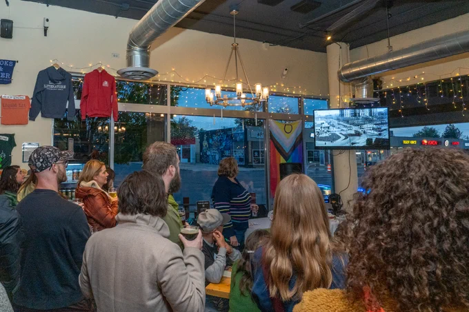 People gathered in a cafe watching the Destination Colfax trailer on a small mounted TV