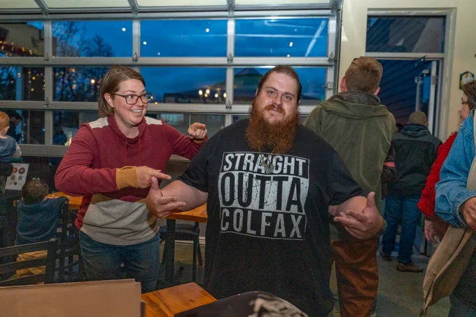 Smiling woman playfully points at a bearded man wearing a "Straight Outta Colfax" shirt in a crowded indoor space.