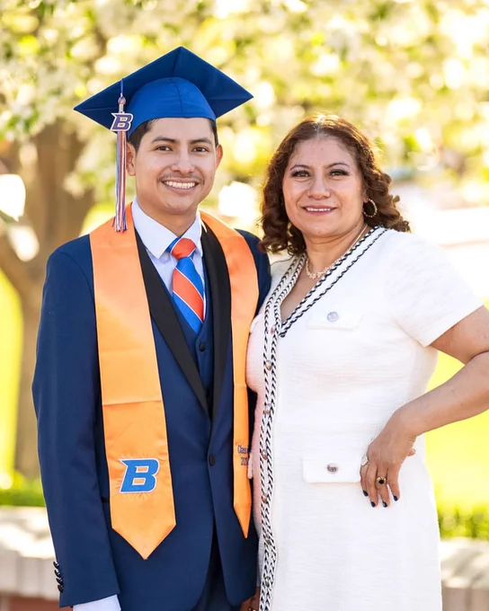 Boise State graduate wearing their stole and cap smiling standing next to their mother. 
