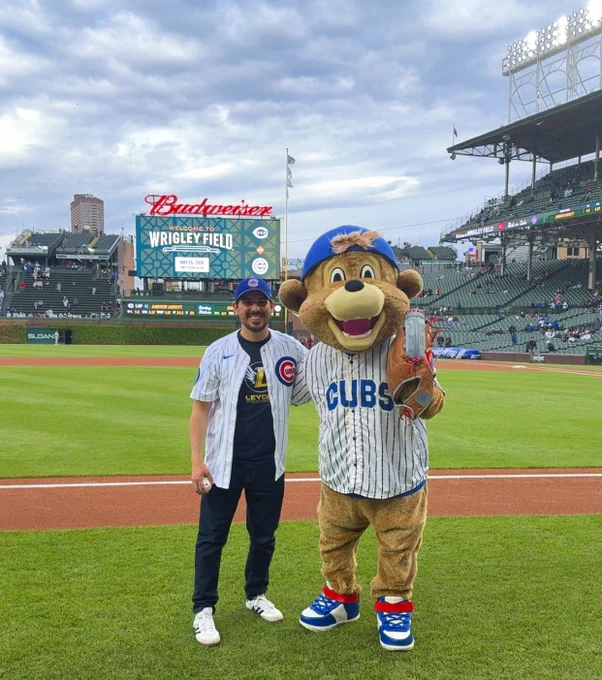 Victor Gomez wears a Cubs jersey and holds a baseball while standing on the field next to the Cubs mascot.