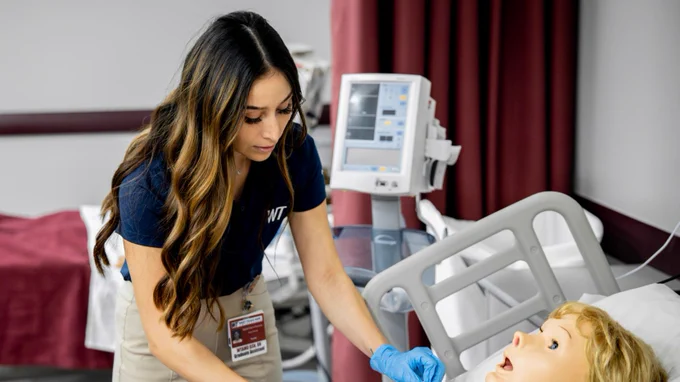 A female with long brown hair and blonde highlights and wearing a navy polo with a WT logo on the left pocket is hovering over a test dummy patient laying in a hospital bed. The room is set up like a hospital room, but is a simulation. The dummy patient has blonde hair.  