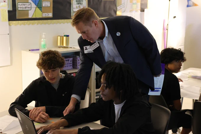Two students are working at a desk, looking at a laptop. Dr. Brad Schnautz is leaning over, pointing at the screen, interacting with the students in a classroom with educational posters and materials in the background.