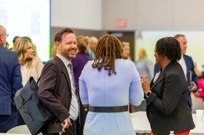 A group of people dressed in professional attire is gathered in an ESC Region 11 conference room, engaged in conversation. There's a presentation screen in the background. 