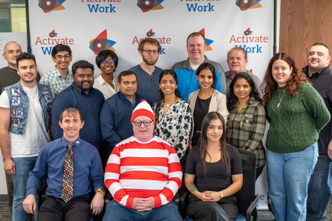 Group photo of ActivateWork's inaugural Data Analytics cohort posing in front of an ActivateWork banner at their graduation ceremony. Seated front and center is John Tillett, smiling in his "Where's Waldo?" costume.