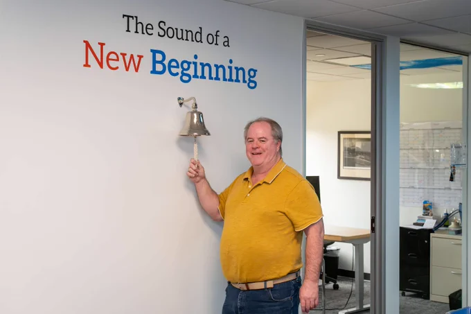 Man in a yellow shirt ringing a silver bell mounted on a white wall with the phrase 'The Sound of a New Beginning' above it.