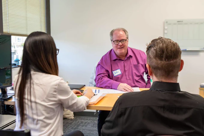 Two interviewees facing volunteer interviewer John Tillett, seated across a table in a professional office setting.