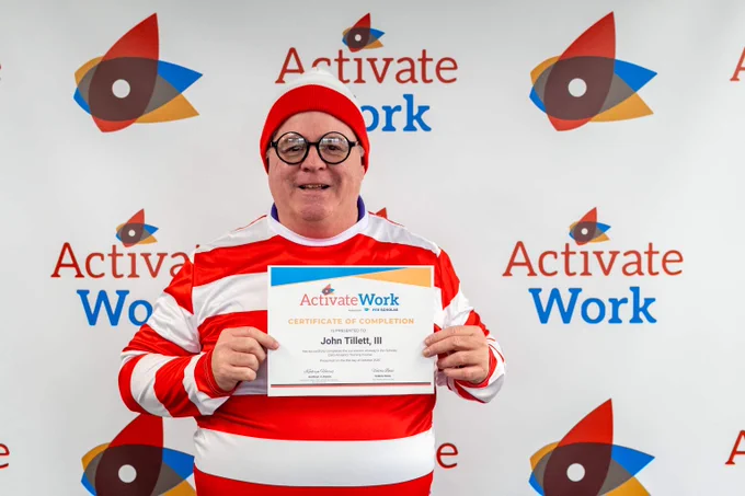 Man in a "Where's Waldo?" costume, holding a certificate of completion from ActivateWork against a branded backdrop.