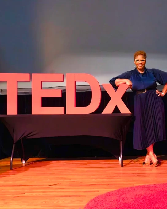 A person stands confidently on a TEDx stage next to large red letters. The environment suggests a formal and inspiring setting.