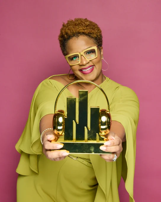 A joyful woman, dressed in lime green, holding a shiny gold award in front of a pink backdrop. Her confident pose and bright smile convey a mood of celebration and achievement.