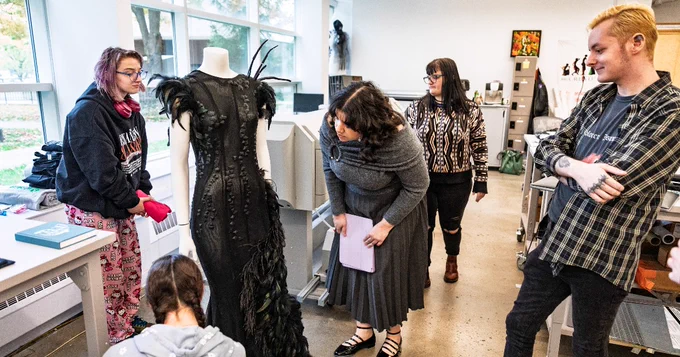 A group of people in a design studio admire an elegant black dress with feather details on a mannequin. The room is spacious with large windows and various design materials scattered around.