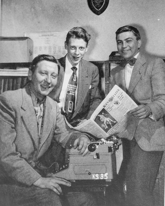 Three people are gathered around a typewriter. One is seated, holding papers and smiling. Another person is standing and also smiling, holding a newspaper called "Daily Kent Stater" The walls have various documents and a shield-shaped plaque.