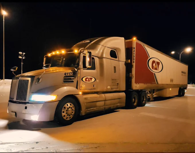 White C.A.T. inc semi truck with a long trailer parked at night in a snowy lot in Calgary, illuminated by warm streetlights, with fresh snow covering the ground and truck tires partially embedded in snow.