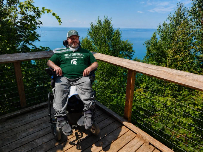 Mo Gerhardt in a wheelchair wearing a green T-shirt with "Built Different: 100% Every Spartan" on the front while on a wooden overlook by a large blue lake.
