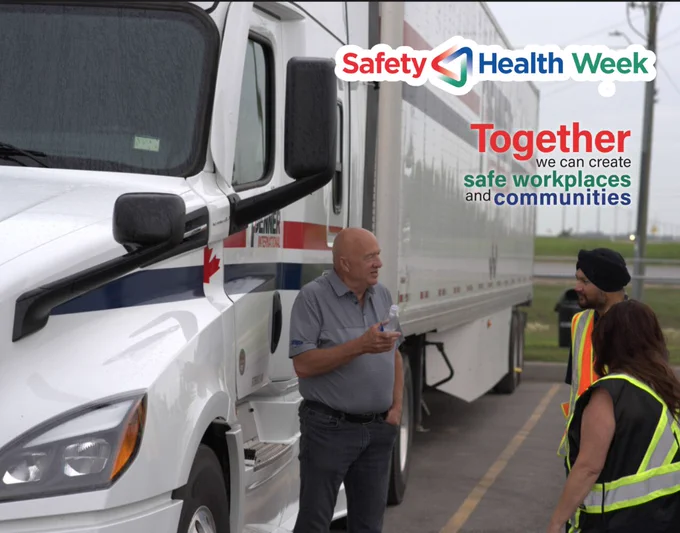 Three workers stand beside a white transport truck with a trailer in a parking area, having a conversation. Two workers are wearing high-visibility safety vests. The truck displays a red maple leaf and company branding. Overlaid text reads “Safety & Health Week” and “Together we can create safe workplaces and communities.” The setting appears overcast, with a roadway and open landscape in the background.