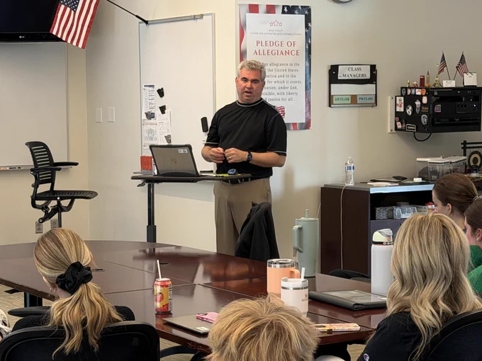 A man in a black shirt standing at a podium speaking to a group of people seated around a table.