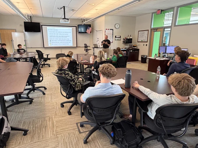 A classroom setting with a group of students seated at desks, facing a large screen displaying text. The room has various learning materials and equipment visible.