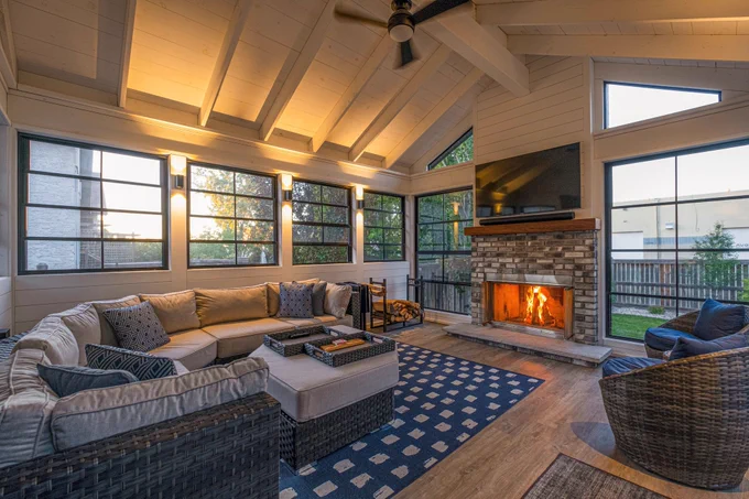 Interior of a four season sunroom featuring large windows, a fireplace, vaulted ceiling with fan, and comfortable seating area with sofa and chairs.