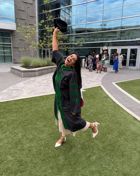 A graduate celebrating outside a modern building with large glass windows, holding up a graduation cap. The individual is dressed in academic regalia and standing on a green lawn, capturing a joyful moment. Other people are gathered in the background near the building entrance.