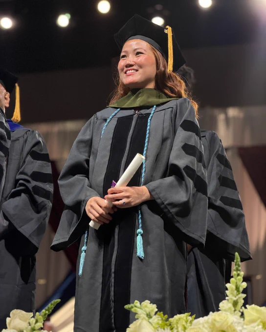 A graduate in a cap and gown stands proudly on stage, holding a diploma. The setting is a formal graduation ceremony with bright stage lights.