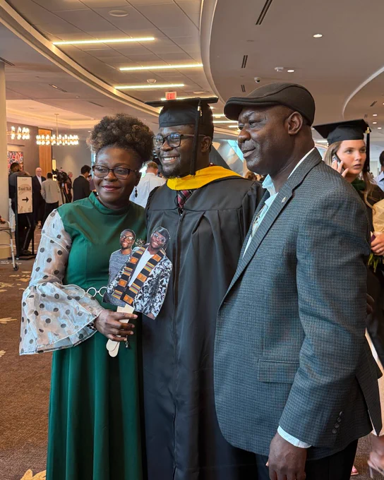 A graduate in a cap and gown holds a cutout image while standing with two people during a ceremony. The interior features elegant lighting and several other individuals in the background.