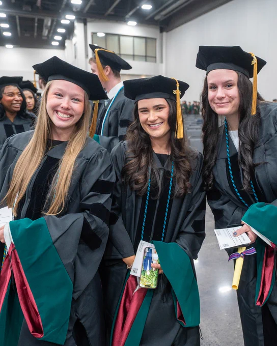 A group of graduates in caps and gowns smile while standing indoors. They hold diplomas and programs, celebrating their graduation.