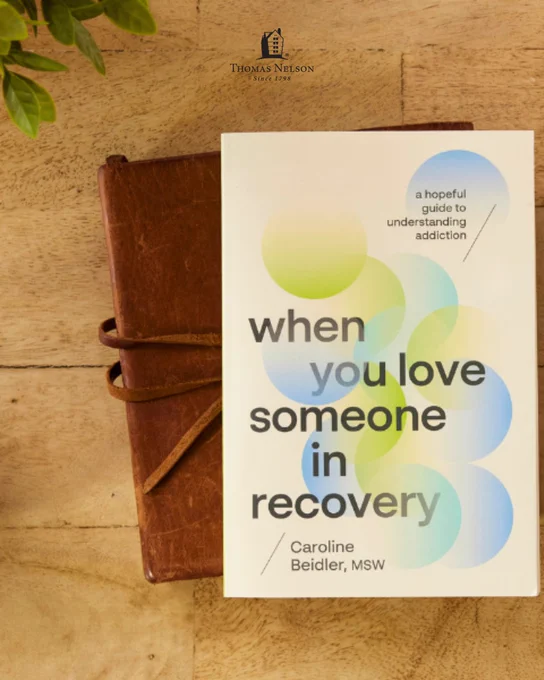 A book titled "When You Love Someone in Recovery" by Caroline Beidler, MSW, rests on a wooden surface with a brown leather-bound notebook underneath. The cover features overlapping blue and green circles with the subtitle, "A hopeful guide to understanding addiction." In the background, there is a small plant and the publisher's logo Thomas Nelson is on the top.
