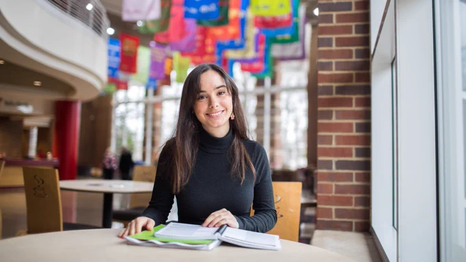 A female student in a black long-sleeved top sits at a table in the Morgan University Center, with bright flags in the background. 