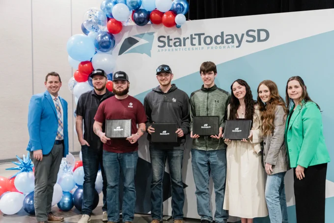 A group of individuals stands in front of a "StartTodaySD Apprenticeship Program" backdrop, smiling and holding certificates. They are surrounded by decorative blue, red, and white balloons.