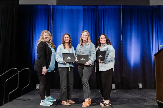 Four individuals stand on a stage in front of blue curtains, each holding a black folder with a logo.