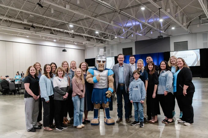 A group of people is standing indoors in a large hall, posing for a photo alongside a mascot dressed in a Spartan costume. There are tables and a projector screen in the background, which displays the text "Northwestern College.