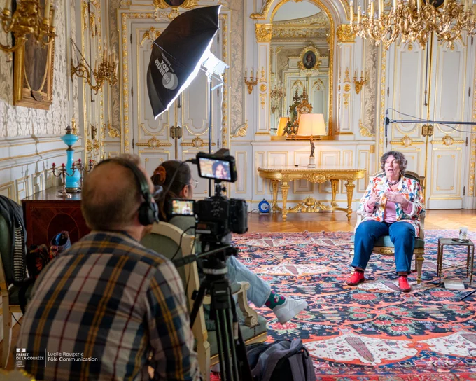 Une femme est interviewée dans un salon luxueux au décor classique et doré, assise sur une chaise devant un tapis coloré. Elle porte une veste à motifs et des chaussures rouges, et s'exprime en regardant la caméra. Face à elle, une équipe de tournage filme avec une caméra sur trépied et utilise un éclairage de studio. Le décor comprend des moulures dorées, un grand miroir, un lustre imposant et des meubles élégants.