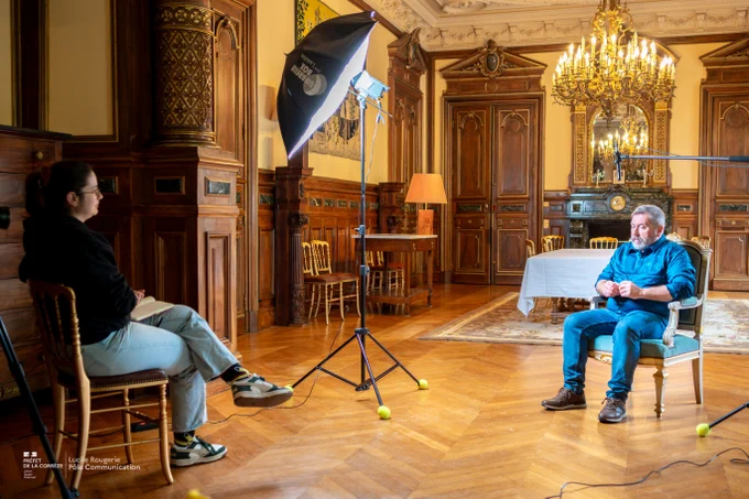 Scène d'interview filmée dans une pièce élégante et ornée de boiseries, avec un lustre imposant au plafond. Un homme assis sur une chaise fait face à une femme prenant des notes, tandis qu'un éclairage professionnel et un micro sont installés entre eux. À l'arrière-plan, on distingue une grande table couverte d'une nappe et plusieurs chaises dorées près de cheminées et miroirs décoratifs.