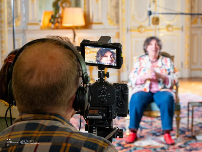 Un cameraman filme une femme assise sur un fauteuil dans une pièce élégamment décorée. La caméra est équipée d’un écran affichant le visage de la femme, tandis qu’elle reste floue à l’arrière-plan. La femme porte une veste colorée et des chaussures rouges. On aperçoit une lampe et des détails dorés sur les murs, typiques d’un intérieur classique. Le cameraman porte un casque audio et une chemise à carreaux.