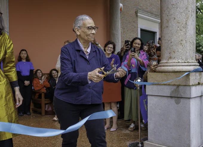 Lubaina Himid cutting a blue ribbon at the opening of the British Pavilion with an audience watching and taking photos.