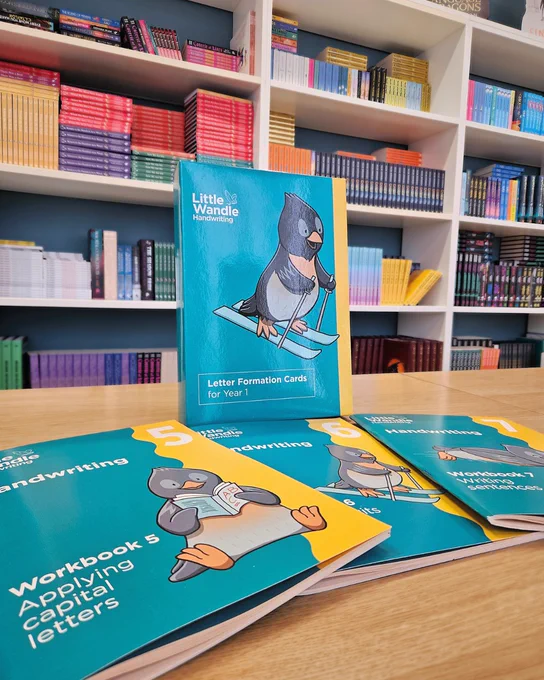 Image of Little Wandle Handwriting workbooks and letter formation cards on a wooden table in front of colourful bookshelves.