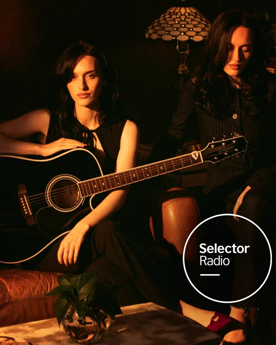 Two sisters, duo Dolder, are dressed in black sit in a dimly lit living room, one holding an acoustic guitar. Selector Radio logo is in the bottom right of the image. 