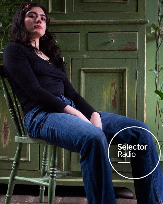Eaves Wilder, a woman with a nose piercing in a black top and jeans reclines coolly on a wooden chair in front of a distressed green cabinet. Selector Radio logo is in the bottom right of the image