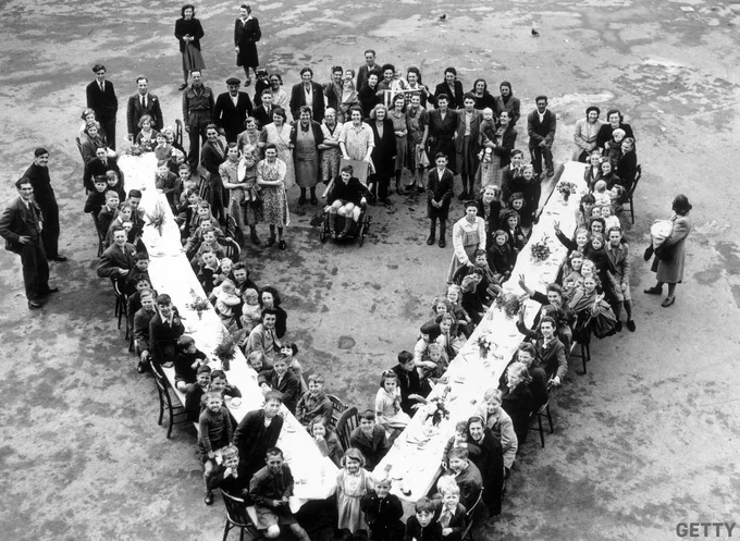 A black-and-white photo of children in London sitting down to a V-shaped table as part of a VE Day party.