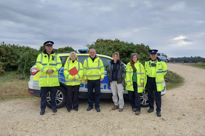 A group of six people stands in front of a marked police car on a gravel car park in the New Forest. They are wearing bright yellow high-visibility jackets. Dense greenery surrounds the area under a cloudy sky.