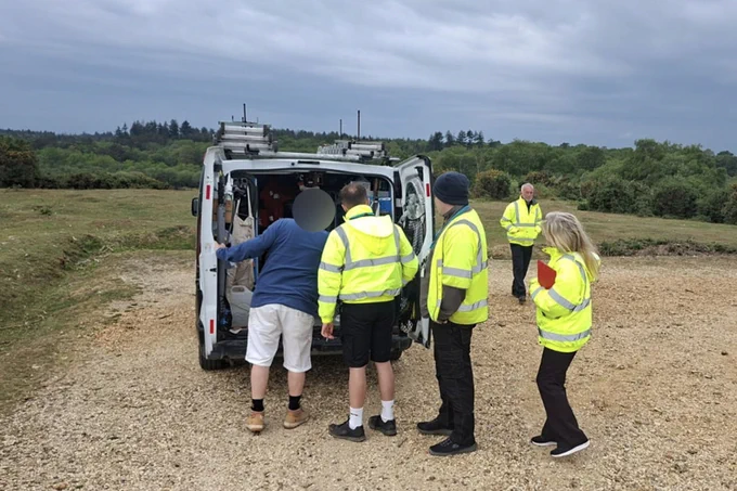 Group of individuals wearing high-visibility jackets stand near an open van in a New Forest car park with trees in the background.