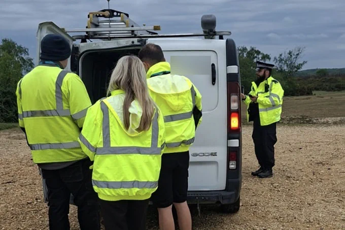 A group of people in high-visibility jackets stand beside a van with its back door open. A police officer is talking to the driver. 