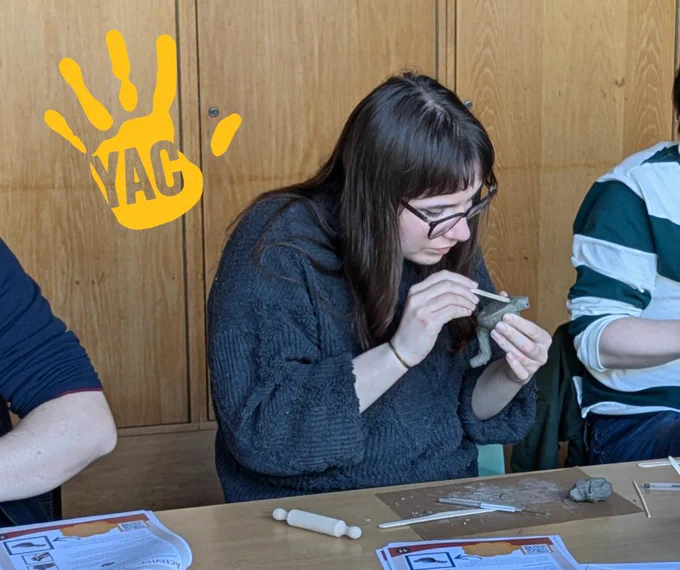 Amelia is sitting at a table and crafting a small object from clay.