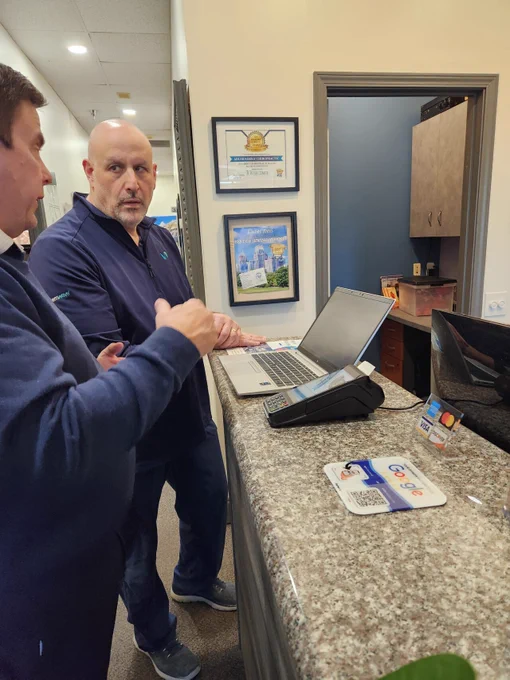 Two men talk at a clinic reception counter beside an open laptop and card payment terminal. Framed awards hang on the wall behind them, and a monitor, card logos, and a Google review QR sign sit on the granite countertop.